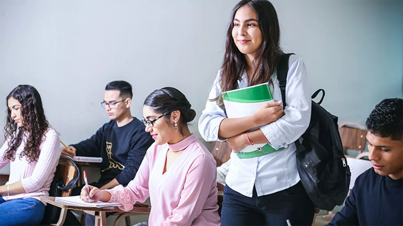 Tjej håller i en bok i ett klassrum, flera personer skriver